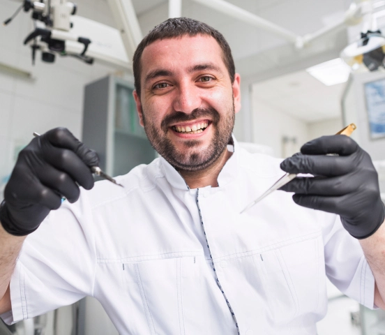 Smiling male dentist wearing black gloves and a white coat holding dental tools in a clinic.
